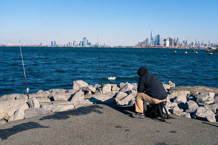 A fisherman sits at the edge of the water with the Manhattan skyline in the distance.